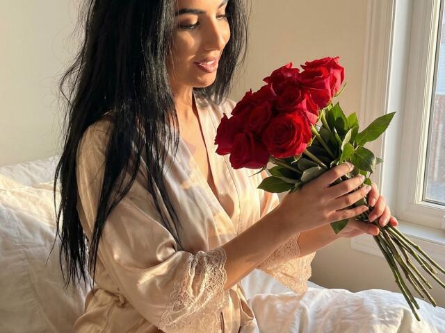 BrunetteBabe69 with long black hair, wearing a beige silk robe, holding a bouquet of red roses, sunlight streaming through a window, soft shadows, slight smile, casual, sensual, intimate setting.