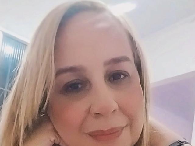 Close-up foto van Remitinha met lichtblond haar, blauwe ogen en subtiele make-up, die lichtjes glimlacht met haar kin rustend op haar hand. aan haar hand. Zacht licht, nonchalante pose, licht blozend gezicht..