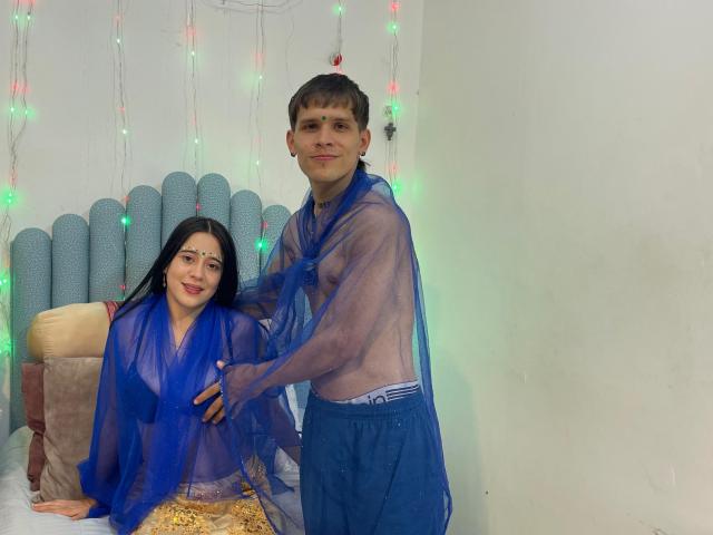 Anjali e Rohan, e um homem em um quarto, ambos vestindo robes azuis transparentes. Ela está ajoelhada em uma cama, ele está atrás dela, com a mão em sua cintura. Luzes de Natal no fundo..