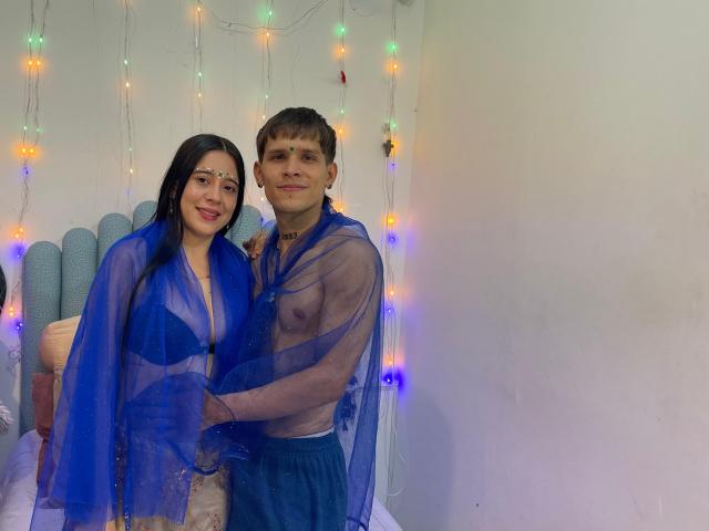 Anjali e Rohan, e un uomo in piedi vicino a loro, entrambi indossano sarong blu trasparenti. Lei ha i capelli lunghi e neri, rossetto rosso e piccoli seni. Lui ha i capelli corti e castani, con un'erezione visibile attraverso il sarong. Luci a stringa multicolore sullo sfondo.