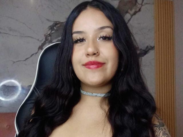 Primer plano de MiaDesiire, una mujer curvilínea con cabello negro largo, lápiz labial rojo y un collar brillante, sentada en un mujer sentada en una silla, mirando a la cámara, sonriendo, con tatuajes visibles en su brazo..