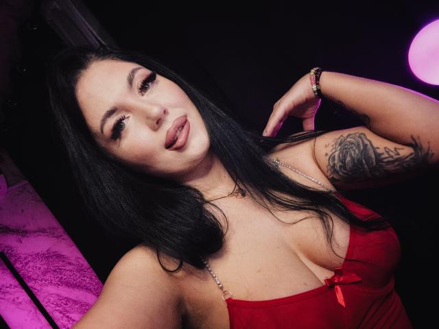 BellaaNoir69, donna dai capelli scuri con un braccio tatuato, che indossa lingerie rossa, con la lingua fuori, sguardo seducente, una mano sul collo. illuminazione viola e rosa, posa provocante, tensione sessuale, sfondo scuro.