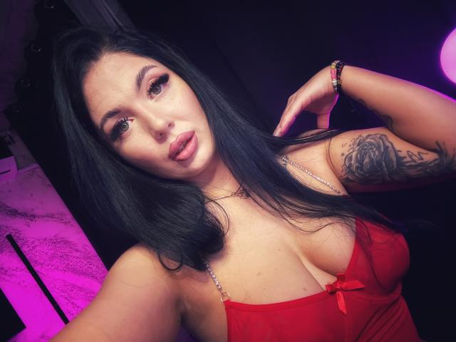 BellaaNoir69, una mujer curvilínea con cabello largo y negro, que lleva una blusa roja de encaje que revela un escote pronunciado. Ella tiene un tatuaje en su brazo derecho y una mirada seductora. Una luz rosa brillante..
