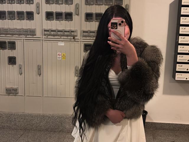 AlessiaNoire, mit ihren langen schwarzen Haaren, macht ein Selfie vor einem Spiegel in einer Umkleidekabine. Sie trägt ein weißes Kleid und einen Pelzmantel und bedeckt ihre Brust mit ihrem Handy. Verdammt heiß..