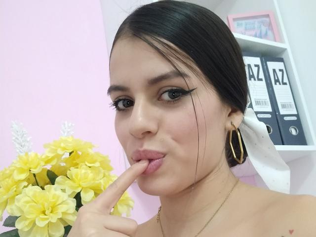 Fotografia di LunaFoxx, una donna attraente con capelli scuri, eyeliner nero e orecchini a cerchio dorati, che si lecca il dito. Mentre teneva in mano un bouquet di fiori gialli, indossando una collana d'oro
.