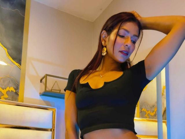 HannaPretty, medium-dark-skinned woman with long brown hair, wearing a black crop top, hand behind head, sexy pose, dimly lit room, gold earrings, soft lighting, subtle blue and yellow accents.