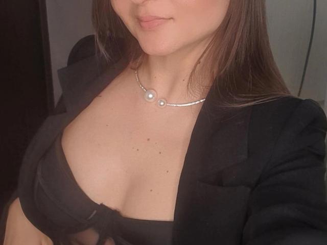 Photo en gros plan de HilyHaraYes, montrant partiellement son visage, avec de longs cheveux bruns, portant un haut noir qui dévoile sa poitrine. et un collier de perles. Elle a une peau claire et un sourire discret. Ses seins sont visibles, avec une légère tension sexuelle..