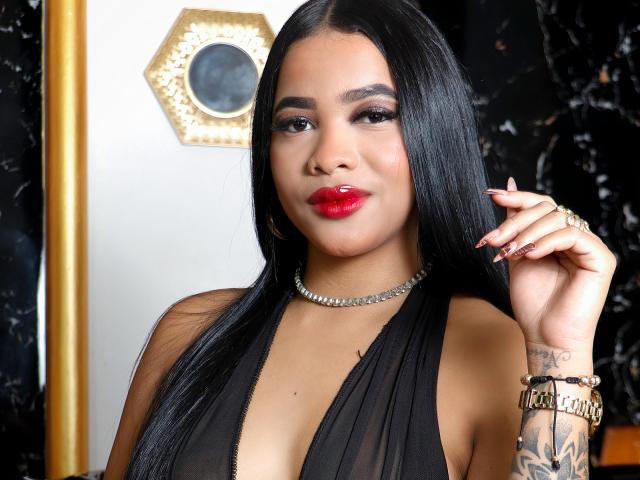 Foto van KarlaBella, een Latina vrouw met lang zwart haar, rode lippenstift en een zwarte jurk met een diep decolleté. Ze draagt een diamanten ketting en armband, en heeft een tatoeage op haar rechterpols. Heel aantrekkelijke, zelfverzekerde pose.