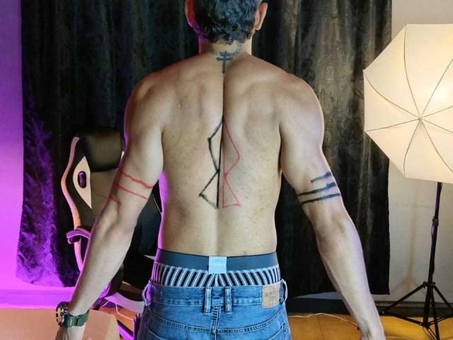 DeconRios senza camicia, con la schiena rivolta verso la telecamera, corporatura muscolosa, capelli ricci neri, tatuaggi tribali rossi e neri sulla schiena, a righe. fasce da braccio, jeans blu, in piedi di fronte a un set fotografico illuminato..