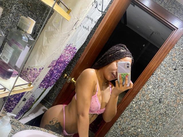 Seductrixz56, com longas tranças pretas, vestindo um sutiã e calcinha de renda rosa, tirando uma selfie no espelho. Tatuagem visível na parte esquerda da nádega, segurando um celular com capa verde. Ambiente de banheiro, toalha roxa..