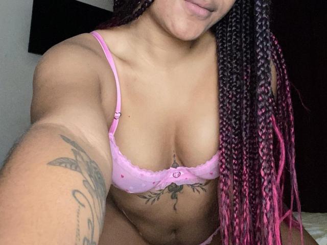 Nahaufnahme eines Fotos von Seductrix56, einer Frau mit gebräunter Haut und geflochtenem, rosa und schwarzem Haar, die einen rosa BH mit einem kleinen Schleifchen trägt. Sichtbare Tätowierungen an Arm und Brust. Verführerische, leicht geöffnete Lippen. Fokus auf Brust und Tattoos.