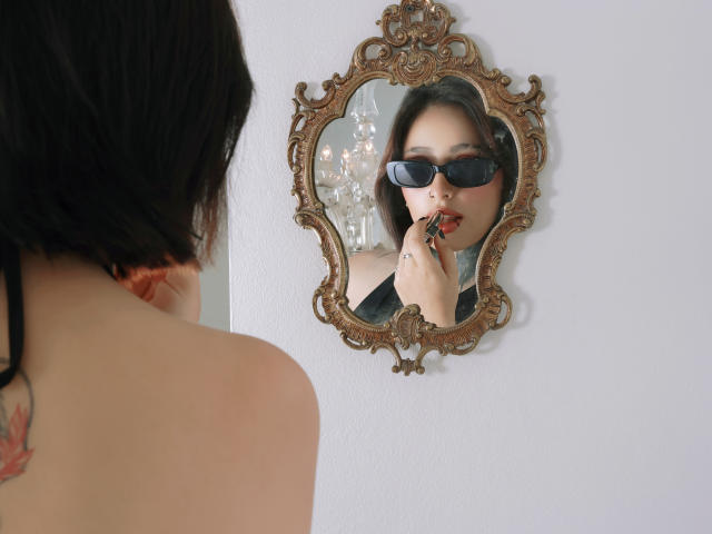 PaisleyMour, com cabelo escuro e batom vermelho, usando óculos de sol pretos, olhando para um espelho dourado ornamentado. Ela está lambendo o dedo, revelando uma tatuagem no pescoço. Uma vibe incrivelmente sexy..