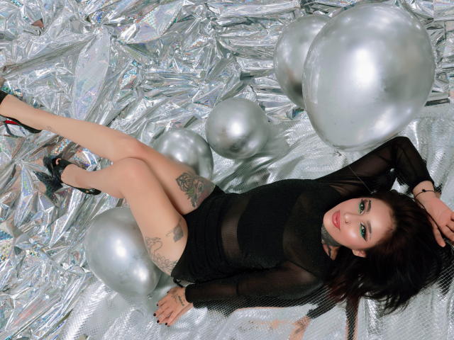 PaisleyMour lies on silver foil with large silver balloons, wearing a sheer black top and black heels. She has tattoos on her arm and thigh, and a sultry expression. Fucking sexy.