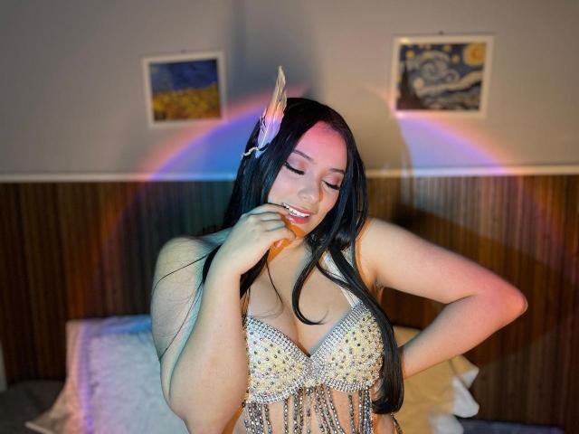 IsabellaCutte with long black hair, wearing a sparkly bra with fringe, touching her lips with a seductive look. A rainbow glow behind her, two framed pictures on the wall.
