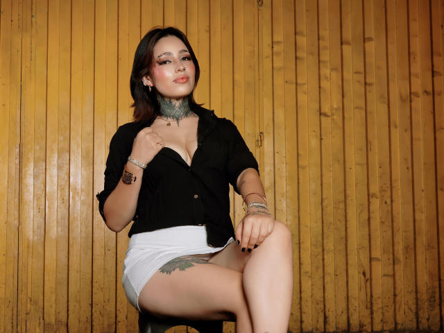 PaisleyMour, con cabello castaño, lápiz labial rojo, camisa negra, pantalones cortos blancos, collar gris y pulseras, sentada con una pierna. de pie, mostrando un tatuaje en su muslo, con un fondo de pared de madera. Increíblemente atractivo..