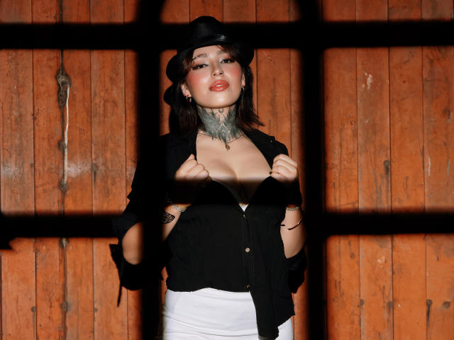 PaisleyMour stands behind a wooden wall with black bars, wearing a black hat and open black shirt revealing cleavage, white skirt, black gloves, and choker. Sexy, provocative pose.