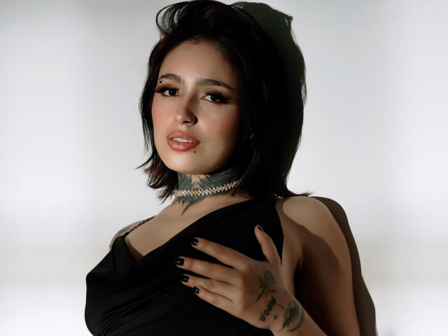 Photograph of a young woman, PaisleyMour, with dark hair, wearing a black top, holding her chest, tattoos on her neck and hand, confident expression, white background, high contrast.