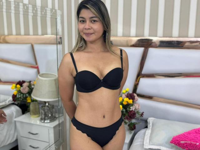 Carly Hamilton steht in einem hell erleuchteten Schlafzimmer und trägt schwarze Dessous mit einem Push-up-BH und Spitzenunterwäsche. Sie hat eine mittelgroße Statur, hellbraune Haut und blondes Haar, das bis zu den Schultern reicht. Bett mit weißen Laken und gelben Blumen im Hintergrund. Die sexuelle Spannung ist in ihrer selbstbewussten Haltung und ihrem leichten Lächeln erkennbar..