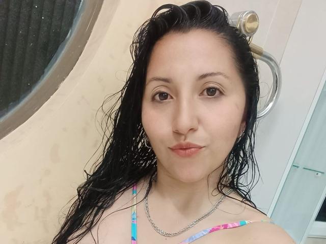 Close-up van AnnittaLatinHot, een vrouw met nat haar, lichte huid en donkere ogen, die een kleurrijke bikini draagt en staat. Voor een spiegel in de badkamer. Ze heeft een subtiele glimlach..