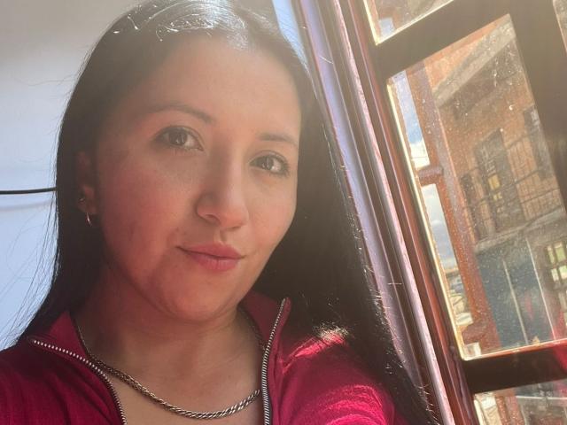 Close-up of AnnittaLatinHot, a woman with long black hair, light skin, and brown eyes, wearing a red zip-up hoodie and silver necklace, standing next to a dirty window, sunlight in background.