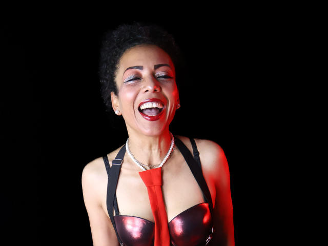 Brenda, met kort, krullend zwart haar, draagt een glanzend zwart leren top en een zilveren ketting, en lacht met rode lippen. rode lippenstift en een brede glimlach. Een felrode das gedrapeerd over haar borst.