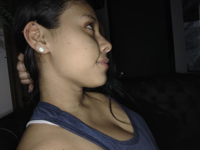 Photograph of MarilynHills, a young woman with medium brown skin, black hair, and a nose ring, wearing a blue tank top, showing side profile, looking away, dark background, earring visible.