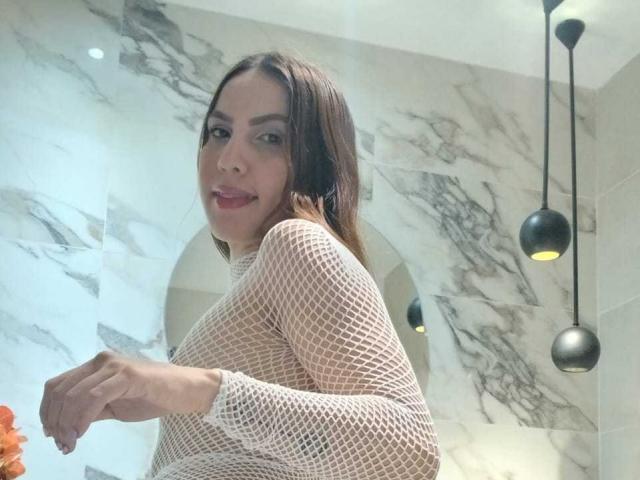SusanThomass, medium-skinned woman with long brown hair, wearing a white mesh top, looking back with a slight smile. Marble wall background, black hanging lights, bright light, subtle sexual tension.