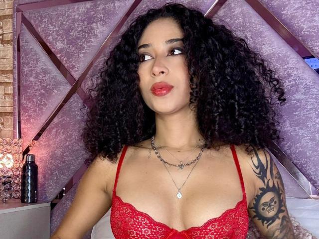Photograph of LannaJhonson with curly black hair, red lace bra, and tattooed arm, gazing upwards with red lips. Dimly lit bedroom with geometric wall pattern and a black bottle. Fucking sexy.