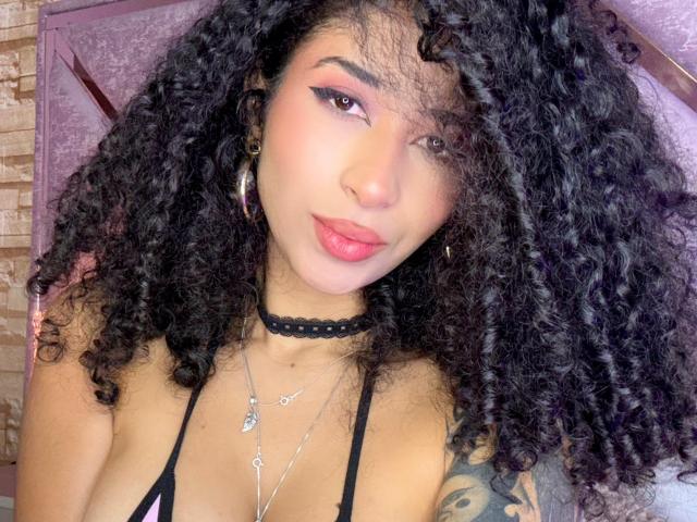 Close-up photo of LannaJhonson, a young woman with long, curly black hair, wearing a black choker and thin-strapped top. She has a tattoo on her right arm and is looking directly at the camera with a slight smile. Her lips are full and pink, and she has hoop earrings. Bright light highlights her face.