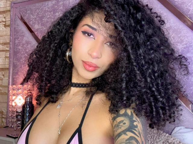Nahaufnahme von Lanna Jhonson mit dicken, lockigen schwarzen Haaren, die einen schwarzen Choker und ein pinkes Bikini-Oberteil trägt und dabei ein Tattoo zeigt. auf ihrer rechten Schulter. Verführerischer Blick, rote Lippen, gedämpftes Licht.