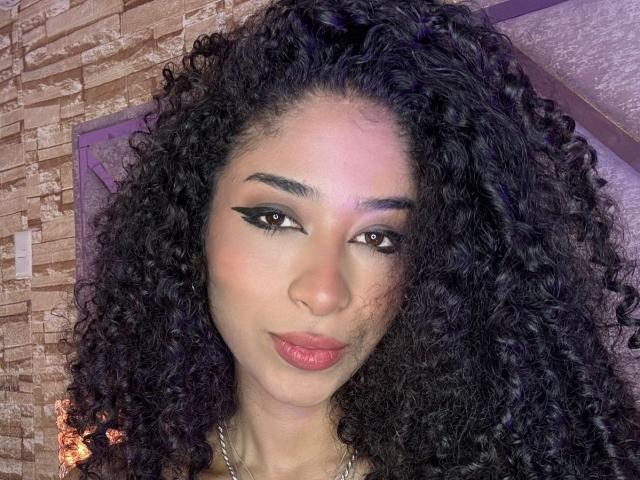 Nahaufnahme von Lanna Jhonson mit langen, lockigen schwarzen Haaren, dunklen Augenbrauen und auffälligem schwarzem Eyeliner. Sie hat eine hellbraune Haut, volle Lippen mit rosafarbenem Lippenstift und ein kaum merkliches Lächeln. Der Hintergrund hat eine Ziegelstruktur..