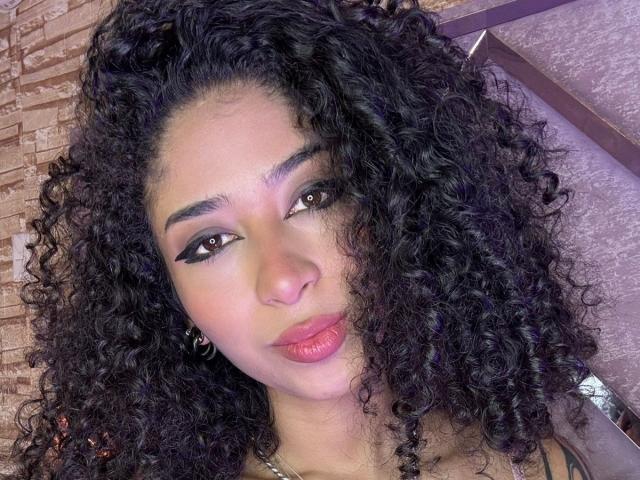 Close-up of LannaJhonson with long, curly black hair, light brown skin, and full lips slightly parted. She gazes directly at the camera with dark, almond-shaped eyes. Her expression is sultry.