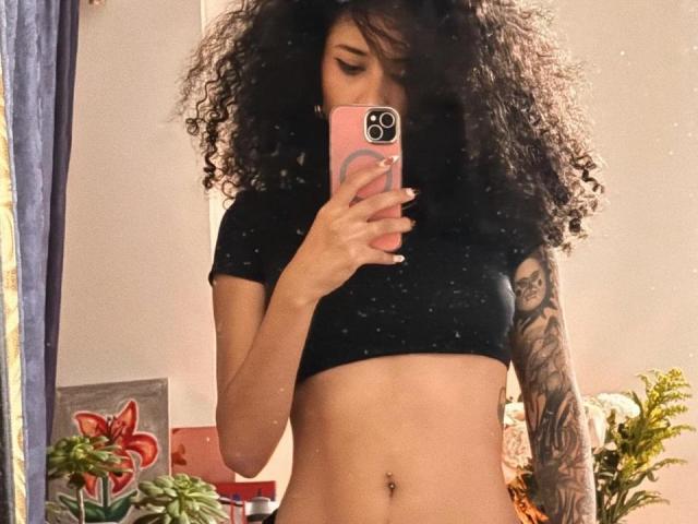 Lanna Jhonson si scatta un selfie allo specchio con un telefono rosa, coprendo il viso con i suoi riccioli neri. Indossa un top corto nero, che lascia scoperto il ventre e mostra il braccio tatuato. Molto attraente..