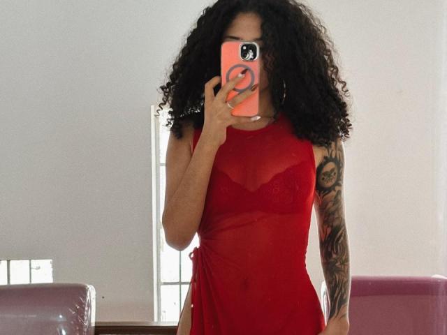 Lanna Jhonson, mit lockigem, schwarzem Haar, trägt ein durchsichtiges, rotes Kleid und hält ein orangefarbenes Telefon mit einem schwarzen Ring. Sie hat Tätowierungen am Arm. Vor einem hellen Fenster stehend, sexuelle Spannung..