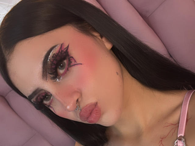 Close-up of LunaaBlossom with black hair, pink blush, heavy eyeliner, and a nose ring. She pouts her lips, showing a pink collar. Her eyes are wide and intense. Fucking sexy.