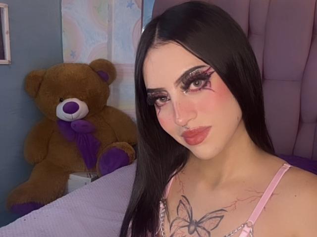 LunaaBlossom, avec ses longs cheveux noirs, son maquillage prononcé et son tatouage de papillon sur la poitrine, porte un top rose à bretelles fines. En haut, elle est assise à côté d'un grand ours en peluche marron. Un look incroyablement séduisant..