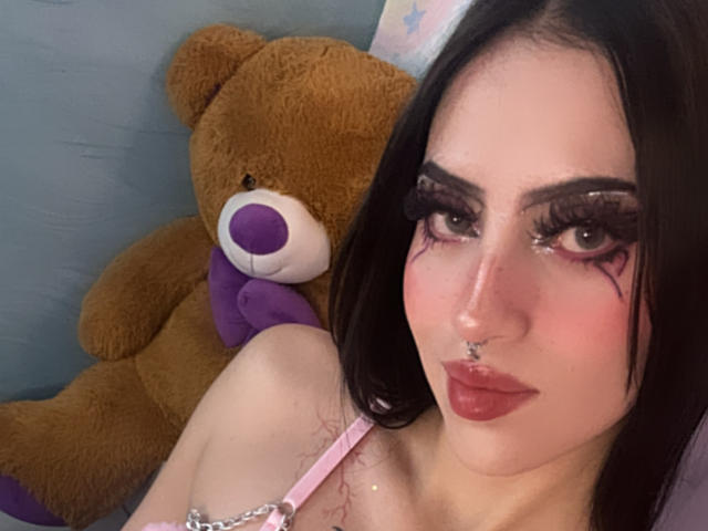 LunaaBlossom with pale skin and dark hair, heavy black eyeliner, and a nose ring, leans against a brown teddy bear with a purple nose, wearing a pink strap. Fucking sexy look.