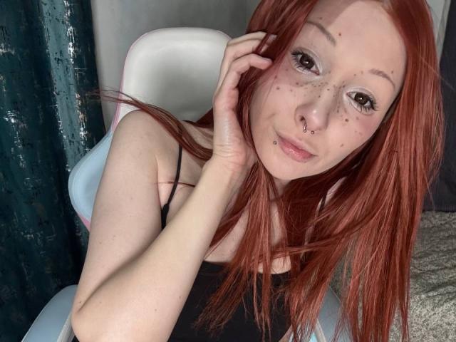 Close-up of HannaCool, a fair-skinned redhead with freckles, piercing nose ring, and long hair, leaning forward on a white chair, hand near face, subtle sexual tension, soft light.