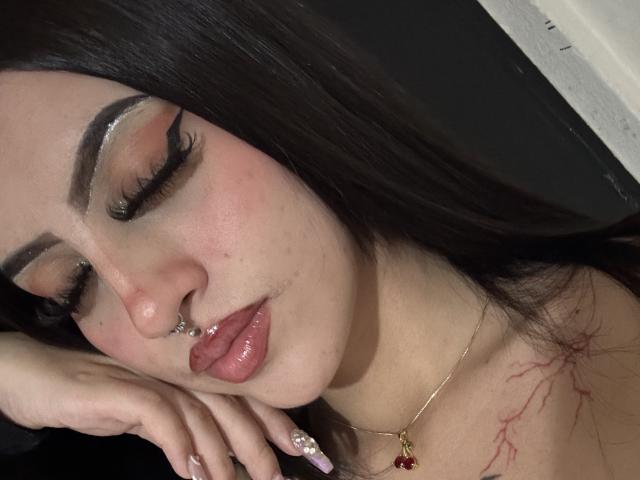 Photo en gros plan de LunaaBlossom, avec de longs cheveux noirs, un trait d'eye-liner prononcé et des lèvres roses brillantes. Elle dort, une main sous le menton, et porte un collier en or. Cou de nuque marqué, visible. Incroyablement sexy..