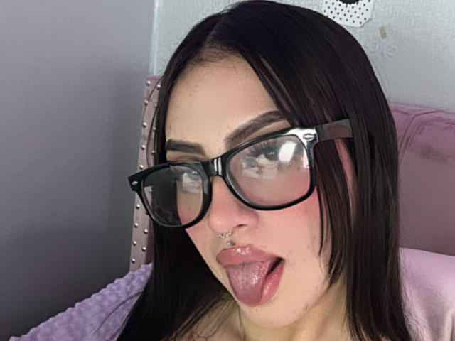 Close-up foto van LunaaBlossom, een jonge vrouw met bleke huid, zwart haar en grote zwarte monturen. Ze steekt haar tong uit, waarbij een lip-piercing zichtbaar is. Op de achtergrond is een hoofdbord met studs te zien..