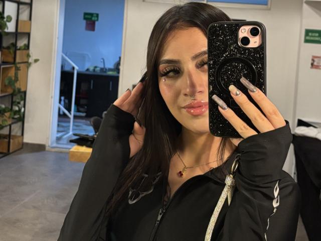 LunaaBlossom, dark-haired, holding a sparkly black phone, wearing a black hoodie with white accents, nose ring, and a gold necklace. Hands in hair, slight smile, office background, sexual tension.