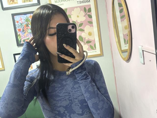 LunaaBlossom mit langen, dunklen Haaren, trägt einen blauen Pullover mit Muster und macht ein Selfie mit einem schwarzen Handy, die Hand ist im Bild. Haare, Augen geschlossen, trägt ein goldenes Armband, sexuelle Spannung ist erkennbar..