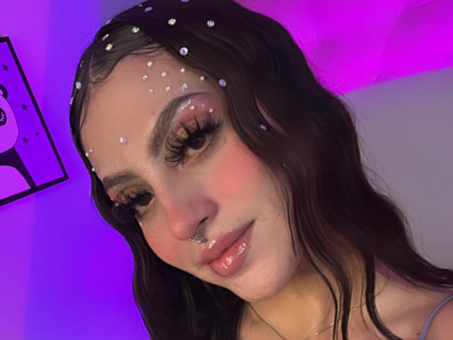 Nahaufnahme von LunaaBlossom mit dunklen, welligen Haaren, verziert mit weißen, sternartigen Punkten auf ihrer Stirn. Sie hat lange Wimpern, rosafarbene Wangen und einen Nasenring. Der Hintergrund ist neonlila und -blau. Verdammt heiß..