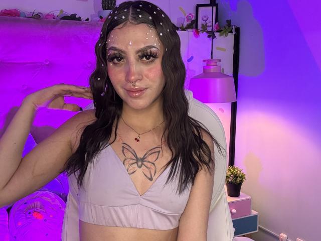 LunaaBlossom, uma mulher com cabelo preto molhado, top cropped branco e tatuagem de borboleta, posa de forma sedutora. Ela está usando glitter no rosto e um piercing no nariz, com iluminação roxa e azul ao fundo..