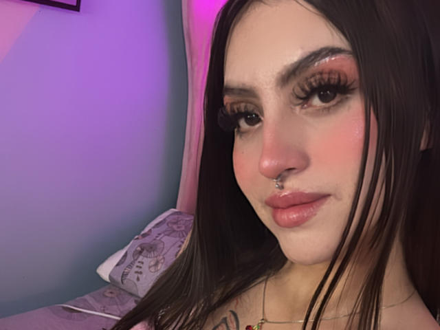 Close-up de LunaaBlossom com cabelo castanho escuro, delineador marcante e cílios longos. Ela tem um piercing no nariz e lábios cheios. Uma iluminação suave em tons de roxo e rosa realça seu rosto. Muito atraente..