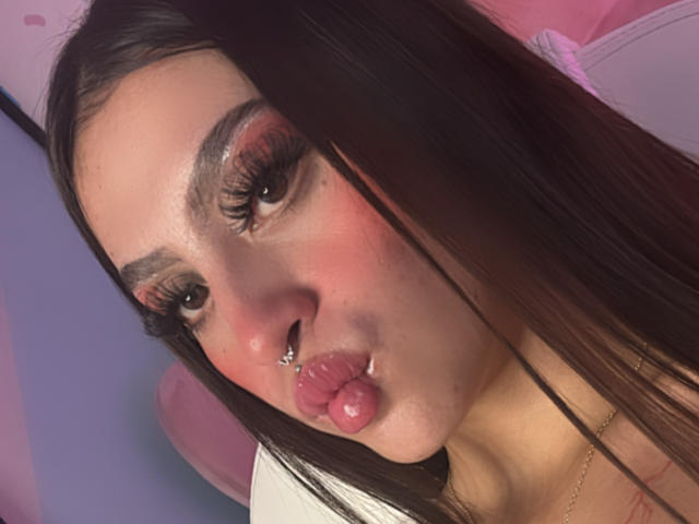 Close-up of LunaaBlossom, pouting with glossy pink lips, dark eyebrows, heavy eyelashes, and a nose ring. Her skin is fair with a slight blush. Long dark hair partially covers her face.