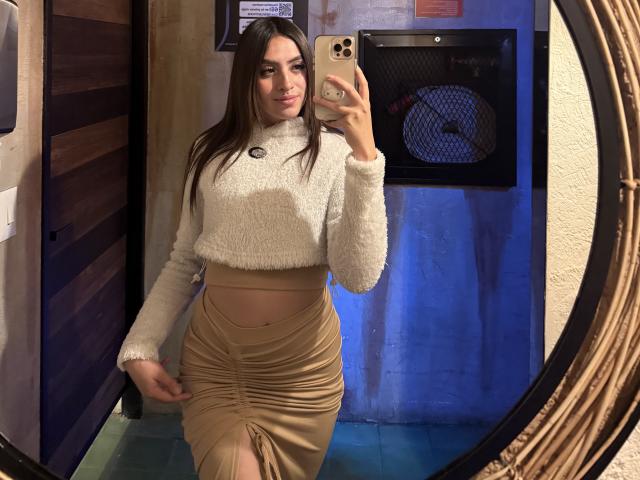 LunaaBlossom, una mujer de cabello oscuro, se toma un selfie en el espejo, vistiendo una camiseta corta blanca y esponjosa, una falda beige con fruncidos, y sosteniendo algo dorado. teléfono, pose sensual, fondo azul y marrón, expresión de confianza, estilo casual..