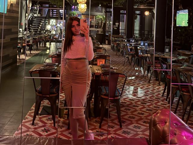 LunaaBlossom se tient dans un restaurant faiblement éclairé et élégant, vêtue d'un haut en dentelle blanche et d'un pantalon moulant à rayures zébrées. Elle pose avec assurance, une main sur sa hanche, entourée de tables vides..