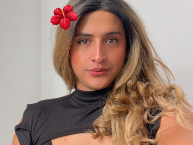 Close-up de LauraCramz com cabelos longos e ondulados loiros, uma flor de hibisco vermelha no cabelo, vestindo uma gola alta preta. Ela tem uma expressão neutra, rosto levemente avermelhado e olhos castanhos..