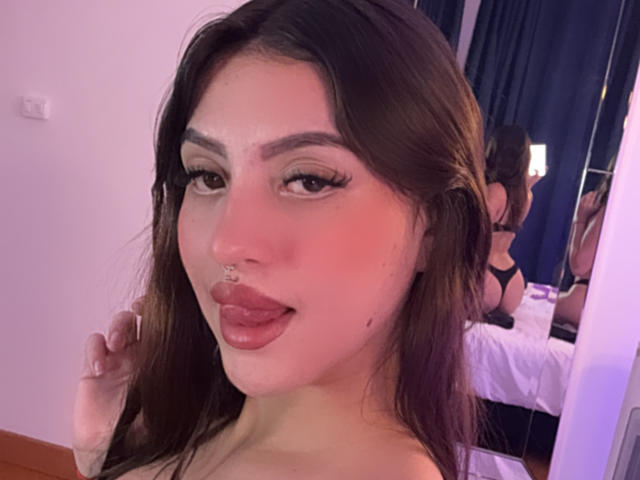 Nahaufnahme von LunaaBlossom, einer attraktiven Frau mit langen braunen Haaren, dicken Augenbrauen und einem Nasenring, die kamera, wobei ihre Zunge leicht herausragt und sie dunklen Lippenstift trägt. Schlafzimmerhintergrund.